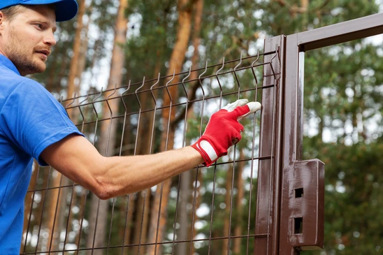 Homme en polo bleu installant une clôture métallique extérieure, portant des gants de protection rouges