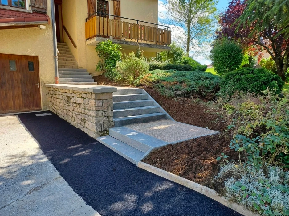 Aménagement extérieur résidentiel : escalier en béton architectonique gris anthracite avec marches bouchardées, mur de soutènement en pierre calcaire apparente, allée en enrobé bitumineux noir