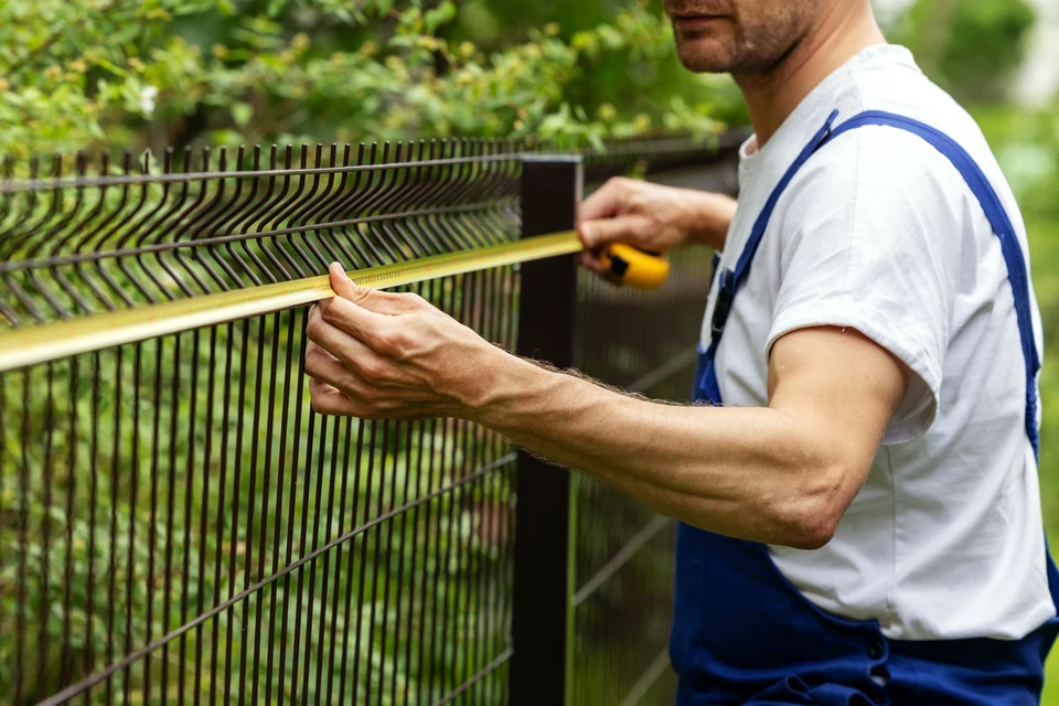 Vue d'un professionnel en tenue de travail mesurant et marquant les points d'ancrage d'une clôture sur un terrain résidentiel, avec un terrain délimité en arrière-plan et les matériaux de clôture (panneaux aluminium ou rigides) visibles sur le chantier. L'image doit montrer le professionnalisme et la précision du travail de préparation d'installation de clôture.